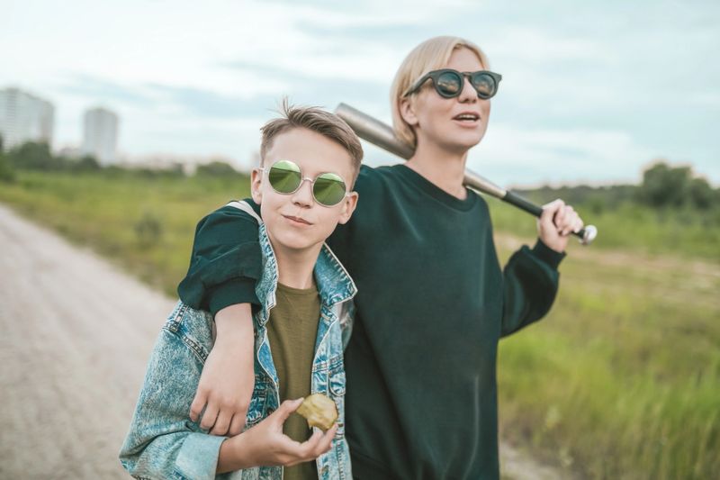 mother and son in sunglasses walking together on ground road, woman holding baseball bat and boy eating apple
