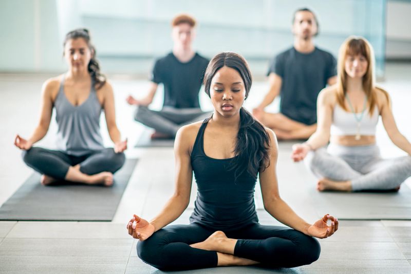 A multi-ethnic group of men and women are indoors in a gym. They are sitting crosslegged and meditating with their eyes closed.