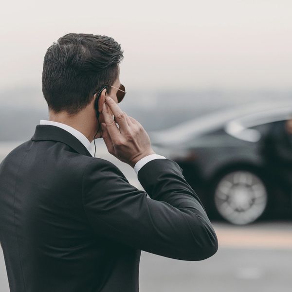 A man in a suit wearing an earpiece stands alert near a black car.