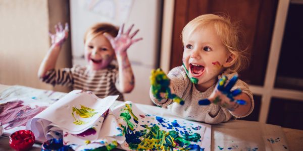 Two young children joyfully painting with colorful handprints indoors.