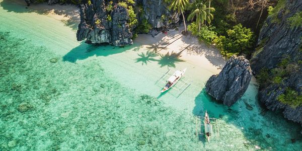 Aerial view of clear turquoise waters, boats, and a small tropical beach with palm trees.