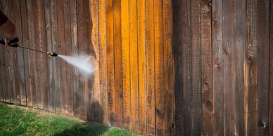 Person pressure washing a wooden fence, revealing a cleaner, lighter section.