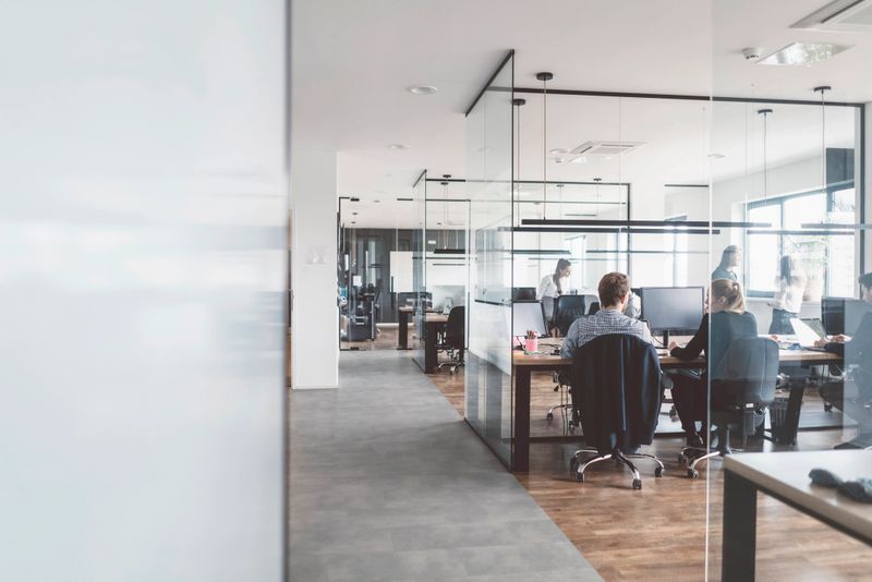 Dark wooden floor and an open space office, a lot of employees working in the office, women wearing business clothes, men wearing elegant shirts and trousers, brainstorming, organising the working process, working on some new projects, glass walls in the office, working on their laptops, working with wireless technology.