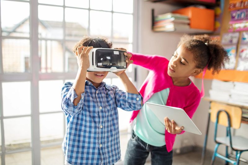 Girl holding digital tablet while assisting boy to wear VR headset. School children using technologies in classroom. They are in elementary school.