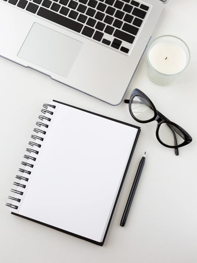 Feminine home office table desk frame. Laptop, notepan and pen, aromatic candle and cat eyewear. Flat lay, top view. Trendy minimalistic concept on the white background. Monochrome colors.