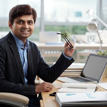 Confident businessman smiling while holding glasses at his desk with laptop and documents.