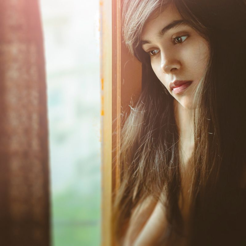 Portrait of Asian, Indian beautiful serene young woman stands near window at home. She looks down and thinks deeply with blank expression.