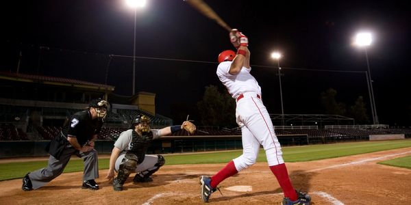 Baseball player swings bat during night game under stadium lights.