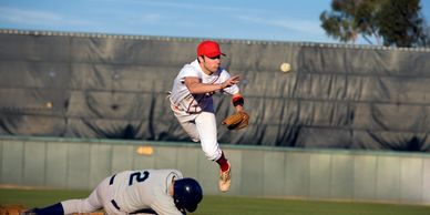 Baseball player leaps over sliding opponent during a game.