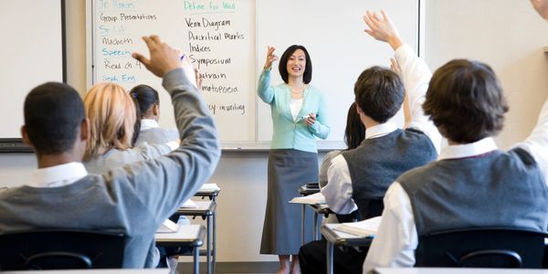 Teacher engaging students in a classroom with raised hands and whiteboard notes.