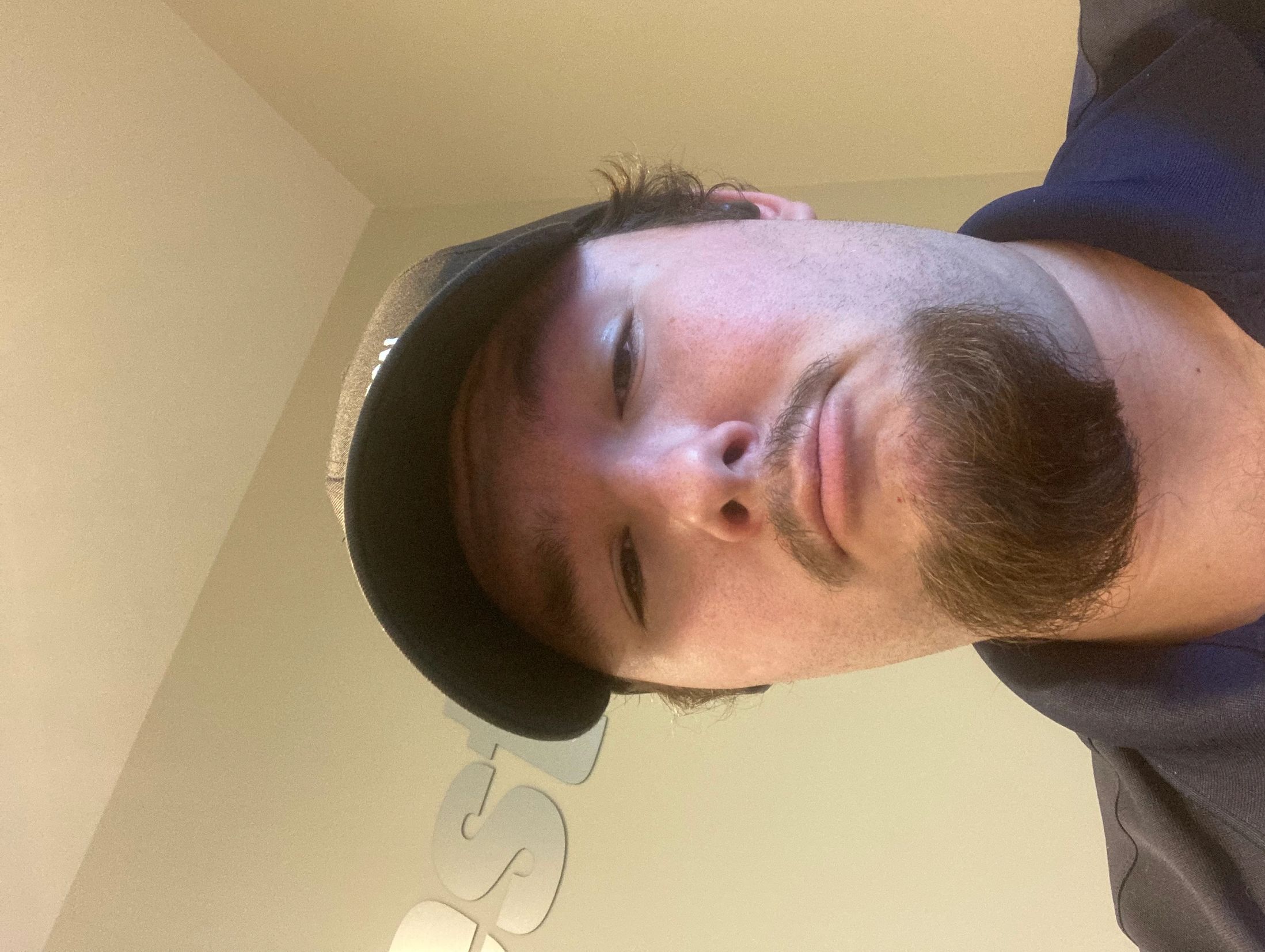 Close-up selfie of a man wearing a black cap and blue shirt indoors.