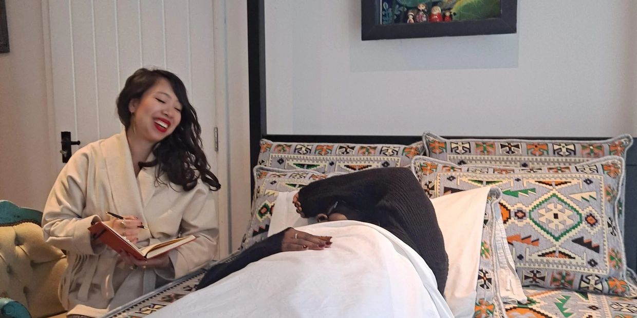 A cheerful woman writes in a notebook while another person rests on a patterned bed.