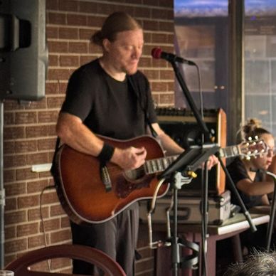Man playing acoustic guitar and singing into microphone indoors.