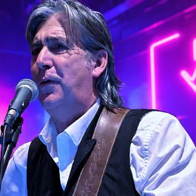 Male musician singing into a microphone on stage with purple lighting.