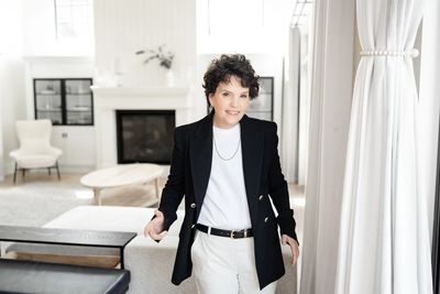 Confident woman in stylish black blazer and white pants in a bright, modern living room.