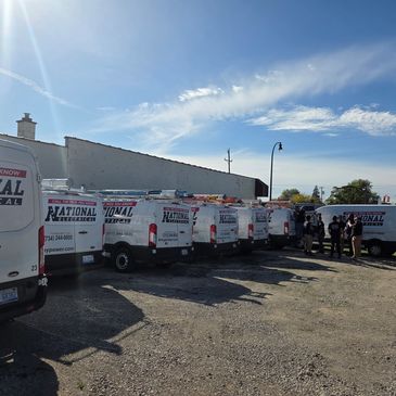 Multiple National Electrical company vans parked with employees standing nearby on a sunny day.