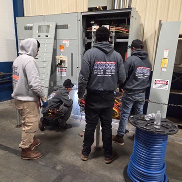 Four electricians working on an electrical panel in an industrial setting.