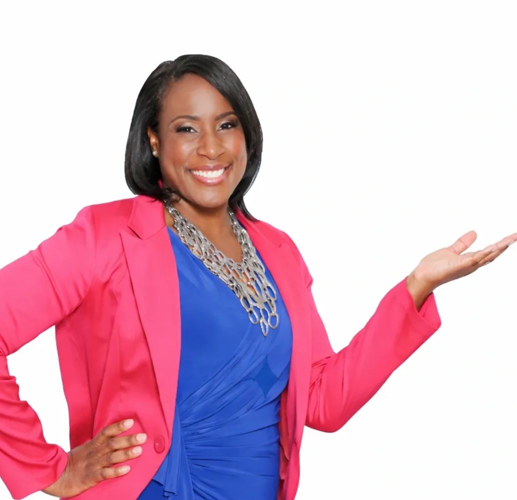 Smiling woman in a pink blazer and blue dress presenting with one hand.