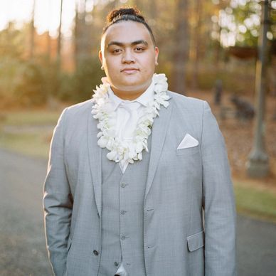 A man in a light gray suit with a white lei and a top knot hairstyle.