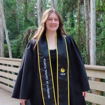 Young woman in graduation gown and stole outdoors near trees.