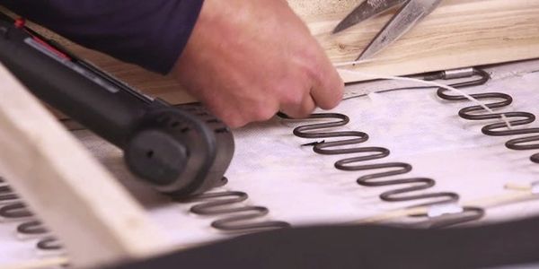 A hand using a staple gun to attach zigzag springs to a wooden frame.