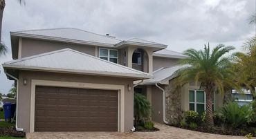 Modern two-story house with a metal roof and palm trees in front.