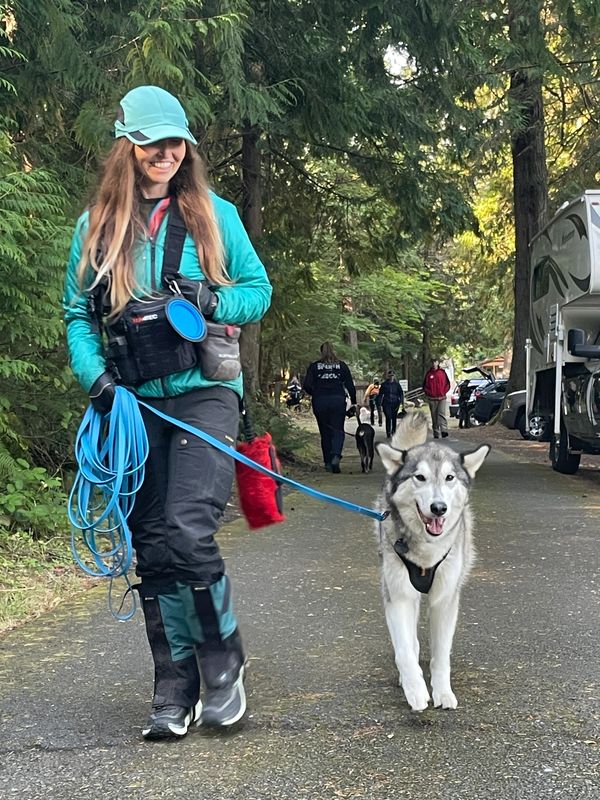 Woman in teal jacket with a husky on a leash in a wooded park.