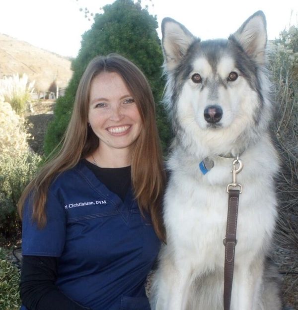 A smiling woman in blue scrubs poses with a large fluffy dog outdoors.