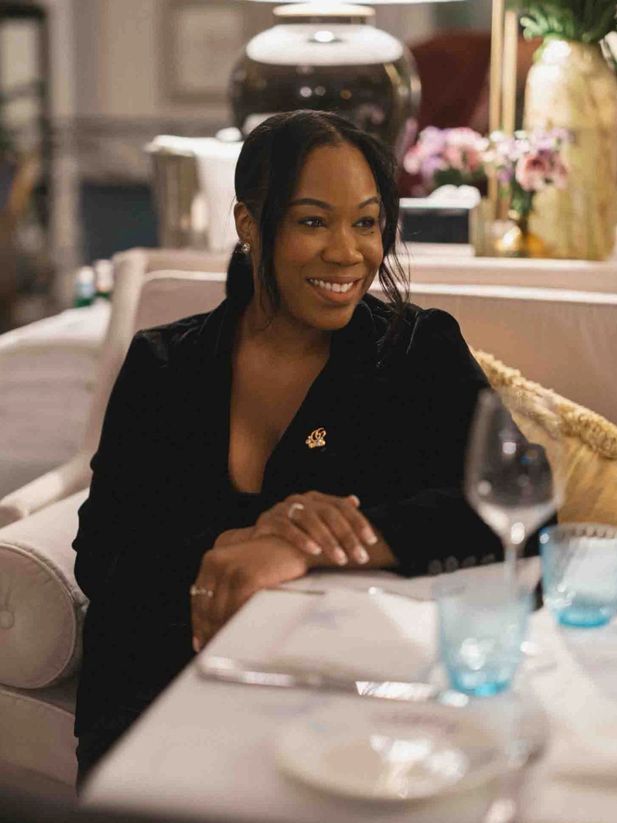 Smiling woman in black blazer sitting at a table in an elegant setting.