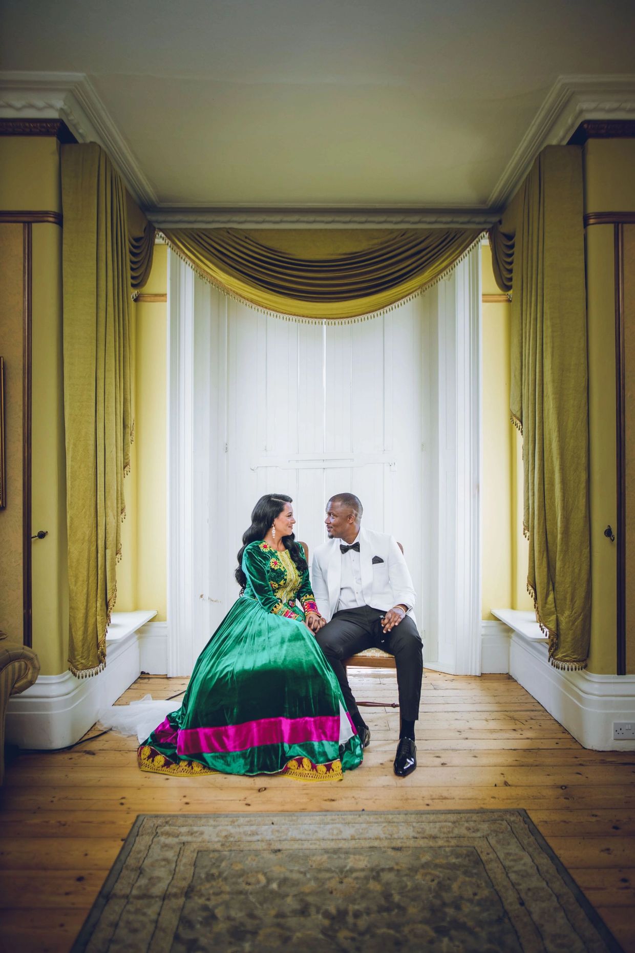 A couple dressed elegantly, sitting and gazing at each other in a bright room.