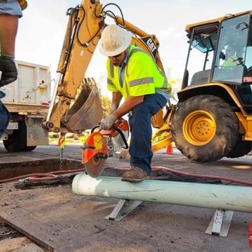 Image of a man cutting a large pipe outside with a tractor and dump truck in the background. 