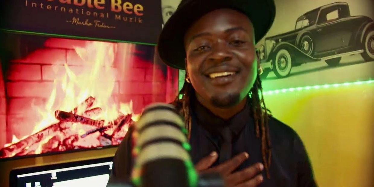 A smiling man with dreadlocks and a hat sings into a microphone in a warm, cozy studio.