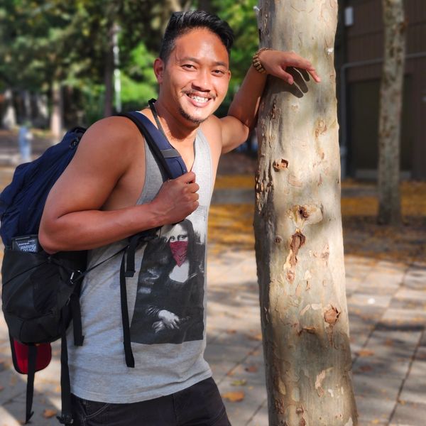 Smiling man with a backpack leaning on a tree outdoors.