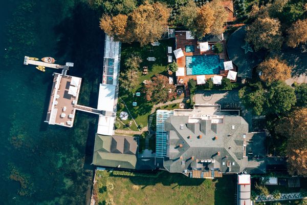 Kuş bakışı otel binası, açık havuz, bahçede şemsiyeler, oturma grupları ve masalar. Göl ve iskele