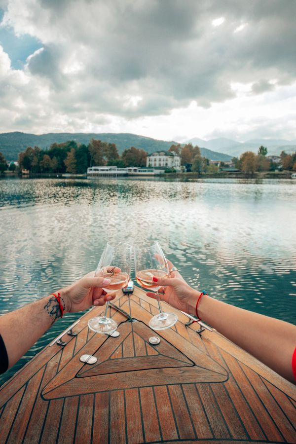 Teknede kadeh tokuşturan iki insan, gölün sakinliği ve ağaçlar arasında kalan otel manzarası