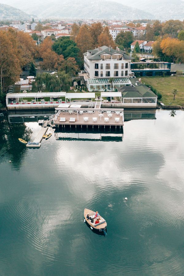 Gölün açıklarında ahşap bir teknede muhabbet eden üç insan, iskele ve ağaçlar arasındaki butik otel