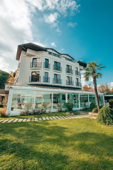 Yeşil bir bahçede, önünde palmiye olan, 3 katlı bir otel binası