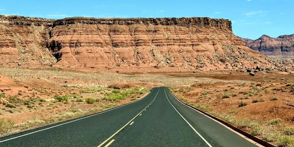 An empty road leads to red rock cliffs under a bright blue sky.