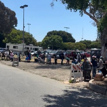 People standing and sitting in a shaded area near parked cars on a sunny day.