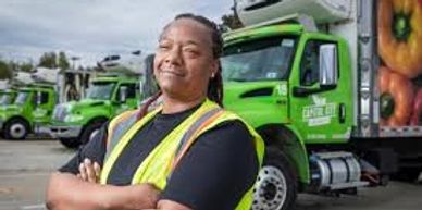 Confident worker in safety vest stands before green delivery trucks.