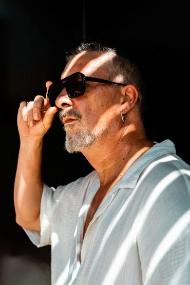 Hombre con gafas de sol y camisa blanca bajo luz de persiana.