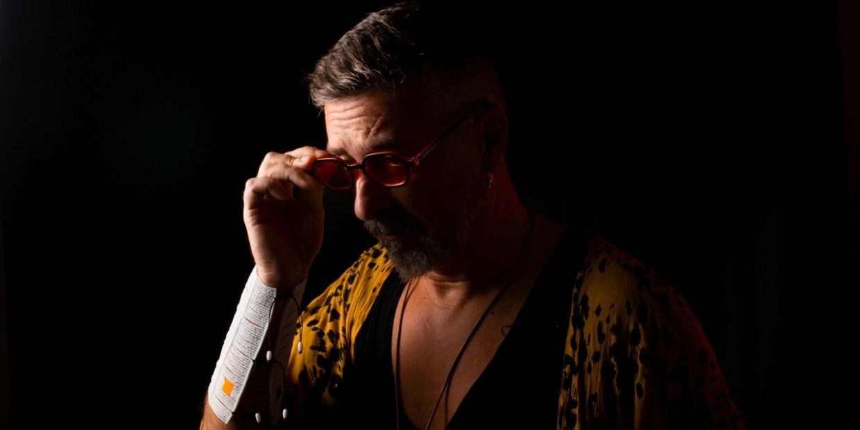 Hombre con gafas rojas y chaqueta amarilla en fondo oscuro.