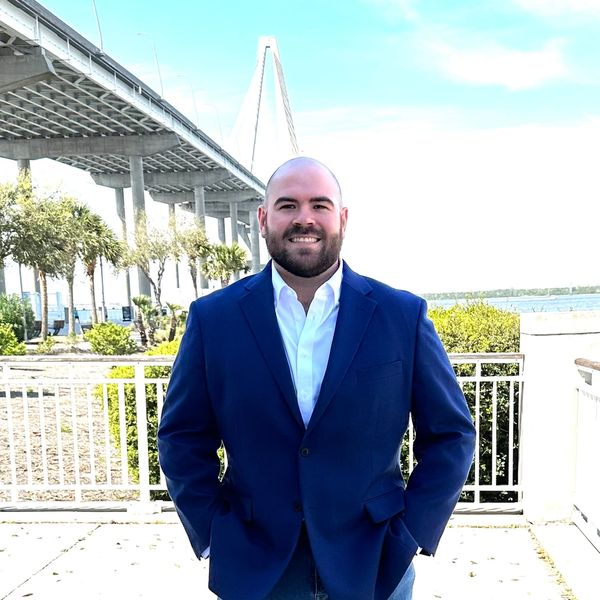 Man in blue suit jacket under a bridge near water.