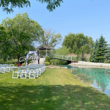 Outdoor wedding setup by a pond with white chairs and a decorated arch.