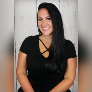 Smiling woman with long black hair in a black top against a white wooden background.
