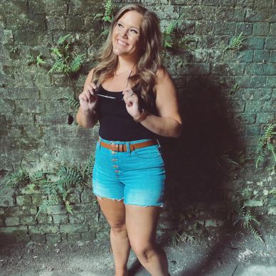 Smiling woman in denim shorts poses against a textured brick wall.