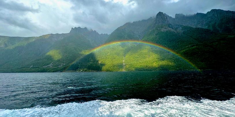 A rainbow arches over a mountainous landscape with water in the foreground.