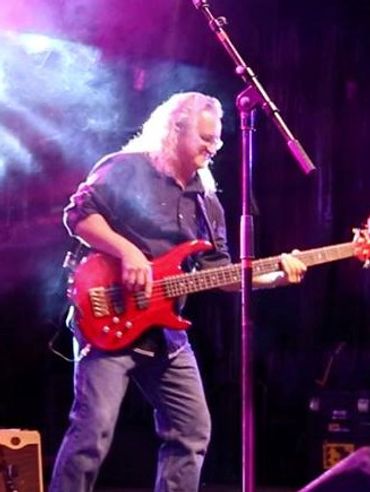 A musician playing red electric bass guitar on stage under purple lighting.