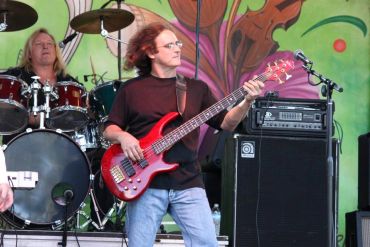 Musicians performing live with bassist in front and drummer in background.