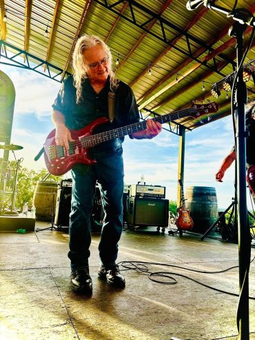Man playing bass guitar on outdoor stage with bright sunlight.